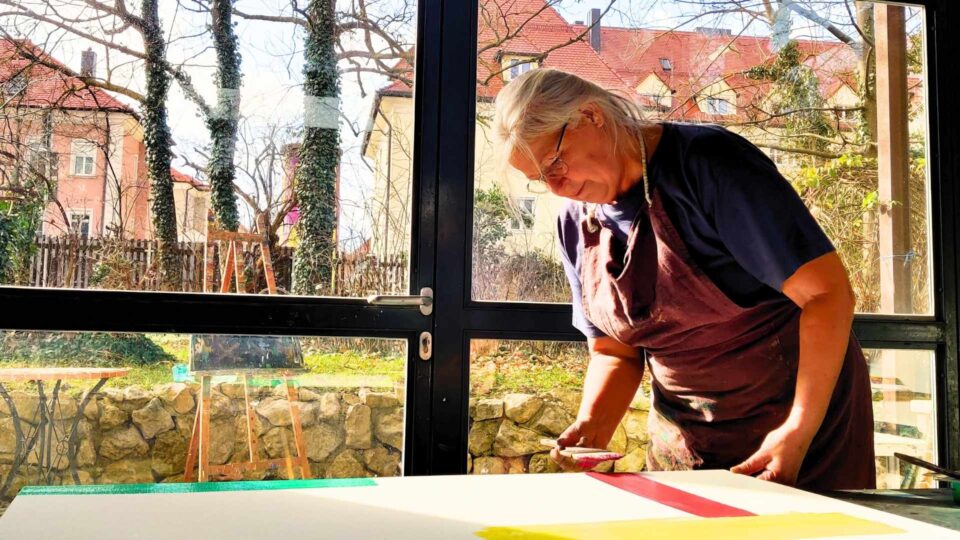 Petra Wild beginnt eine Arbeit in Lasurtechnik und hat einen breiten Pinsel mit roter Farbe in der Hand.
